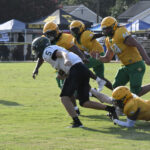 Football players in green and yellow uniforms tackle a player in a white and green uniform during a game on a grassy field.