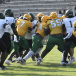 Football players in yellow and green uniforms face off against players in white and green during a game, with empty bleachers visible in the background.