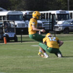 Two football players in green and yellow uniforms, one holding the ball for a kick while the other prepares to kick on a grassy field.