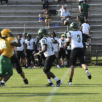 High school football players in green and yellow uniforms play on a grassy field, with teammates and spectators seated in bleachers in the background.