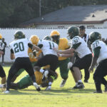 High school football players in green and yellow uniforms tackling and blocking each other on a grassy field during a daytime game.