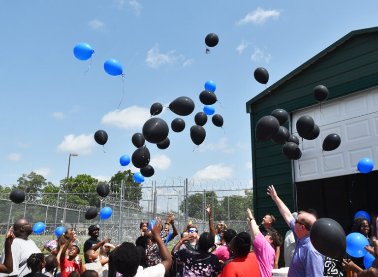 A group of people outside release black and blue balloons into the sky on a sunny day near a green building and a fenced area.
