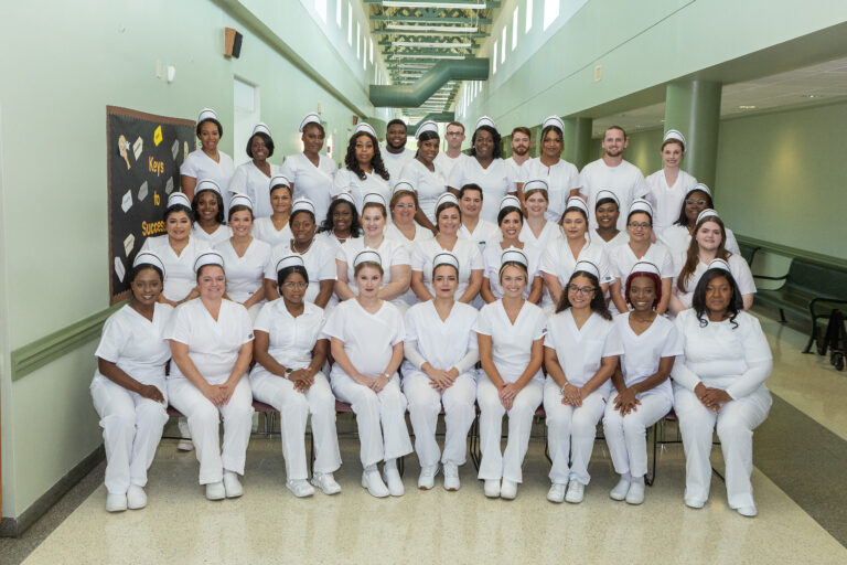 A large group of people dressed in white uniforms pose together indoors, likely students or professionals in a healthcare or nursing program.