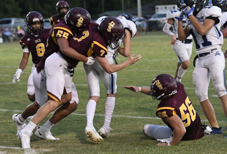 High school football players in maroon uniforms help a teammate up from the grass during a game, with opposing players in white visible in the background.