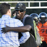 Two men greet each other on a football field, one wearing a cap and sunglasses, while players in orange jerseys stand in the background.