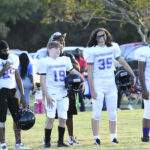A group of young football players in white uniforms stand on a field, holding helmets, with trees and people in the background.