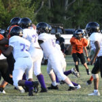 Youth football teams in white and orange uniforms play a game on a grassy field, with players actively engaging in blocking and running.