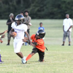 A youth football player in white runs with the ball as an opponent in orange tries to tackle him, with spectators watching in the background.
