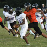 Youth football players in helmets and uniforms run on a grassy field during a game, with one player holding the ball and others in pursuit.