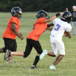 Two young football players in orange jerseys attempt to tackle a player in white jersey number 3 during a game on a grassy field.