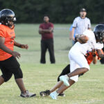 Two youth football players collide during a game, one in white uniform being tackled by another in orange, while referees and a coach watch in the background.