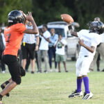 Two youth football players, one in orange and one in white, compete for the ball during a game, with spectators and vehicles in the background.