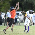Two youth football players in action, one in an orange jersey jumping with arms raised, the other in white preparing to throw the ball, with spectators in the background.