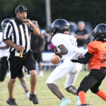 A football player in white runs with the ball while a player in orange tries to tackle him. A referee observes closely on the field.