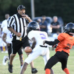 Youth football players in action on a field, referee nearby watching closely, one player in orange and another in white pursuing a loose ball.