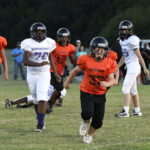 Youth football game with players in orange and white jerseys running on a grassy field, some in pursuit, with trees and spectators in the background.