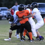 Youth football players in white uniforms tackle a player in an orange jersey labeled “Pirates” during a game on a grassy field.