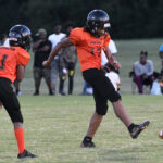 Youth football player in orange jersey kicks the ball during a game, with another player nearby and spectators watching in the background on a grassy field.