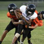Three youth football players in helmets and uniforms during a game, with two players in orange jerseys tackling a player in a white jersey holding the ball.