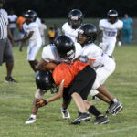 Young football players tackle an opponent in an orange jersey during a youth game, while other players and a referee watch in the background.