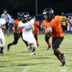A football player in an orange jersey runs with the ball while being chased by opponents in white uniforms during a game.