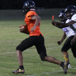 A football player in an orange jersey runs with the ball, chased by two players in white jerseys on a grassy field.