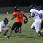 A youth football player in an orange jersey runs with the ball, pursued by three players in white uniforms during a game on a grassy field.