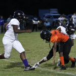 Two youth football players in black helmets collide during a game, one in an orange jersey and the other in white, with another player approaching.