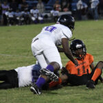 Football players in orange and white uniforms are on the field; one orange player is tackled and on the ground while two white team players defend.