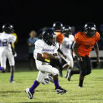 Youth football players in action on a field at night, one player in white running with the ball as others in orange and white chase.