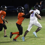 Youth football game at night, player in white runs with the ball while two players in orange jerseys attempt to tackle him on the field.