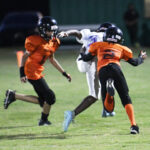 Three youth football players in action on a field at night, two in orange uniforms and one in white, competing intensely during a game.