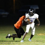 A football player in white runs with the ball while a player in orange attempts to tackle him during a night game.