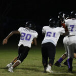 High school football players in white and black uniforms run on a field at night during a game.