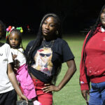Four girls stand together outdoors at night, one holding a small child, all looking toward the camera under artificial lighting on a grassy field.