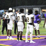 Football players from two teams stand on the field for the coin toss, with referees nearby. The scene takes place on a turf field with purple markings.