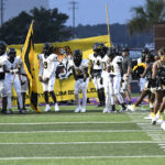 High school football players in white uniforms prepare to enter the field, holding a yellow banner, with cheerleaders standing nearby.