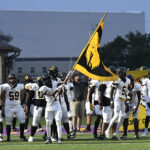 Football players in white uniforms stand on a field, one player holding a large yellow and black flag, with coaches and teammates in the background.
