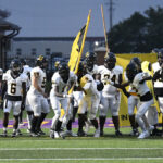 A high school football team in white uniforms runs onto the field carrying yellow flags, preparing for a game at dusk.