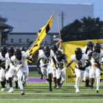 A high school football team in white uniforms runs onto the field, led by a player carrying a yellow and black flag.