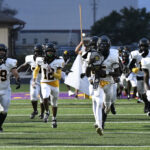 A football team in white uniforms runs onto the field, led by a player carrying a large flag, during the evening.