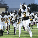 High school football team in white uniforms runs onto the field, with player number 2 leading and cheering, during an evening game.