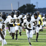 A group of American football players in white uniforms run onto the field during a game, with some players wearing black helmets and yellow gloves.