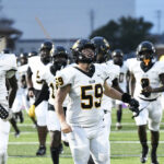 A group of American football players in white uniforms with black helmets walk on a field, with number 59 in front.