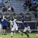 Two high school football players compete for a thrown pass on a field, watched by spectators in the stands.