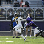 A football player in a white uniform runs with the ball while a defender in purple attempts to tackle him on a field during a game.