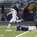 A football player in a white uniform runs past a fallen opponent in a purple uniform on a field, with spectators in the background.