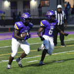 Two football players in purple uniforms run on a field during a game, with a referee and spectators visible in the background.