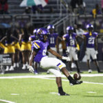 A football player in a purple jersey kicks a football during a game, with teammates and cheerleaders visible in the background on a lit field at night.
