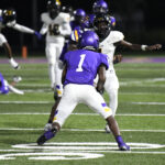Two high school football players facing off during a nighttime game, one in a purple uniform and one in white, on a well-lit field.